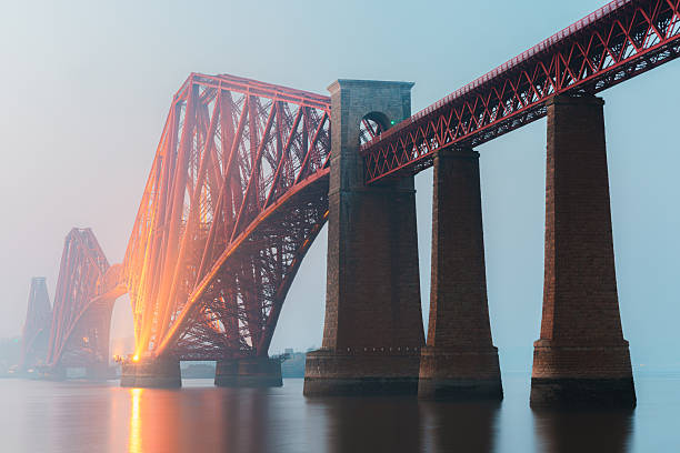 The iconic Forth Bridge spans the calm waters of the Firth of Forth near South Queensferry, Scotland, its red cantilever structure glowing through the  mist during blue hour.