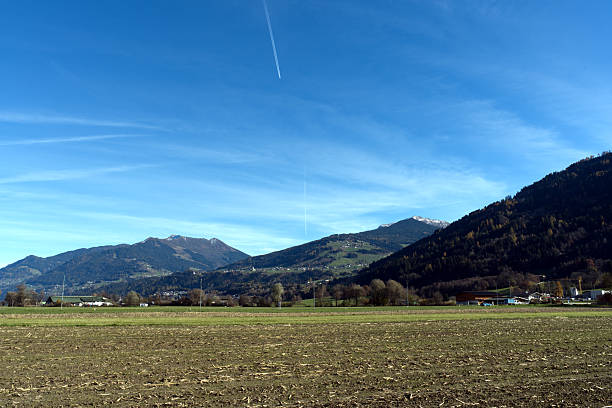 Nestled in a serene alpine valley, this tranquil village scene blends golden fields, forested hills, and snow-capped peaks under a clear sky, evoking harmony between nature and peaceful habitation.