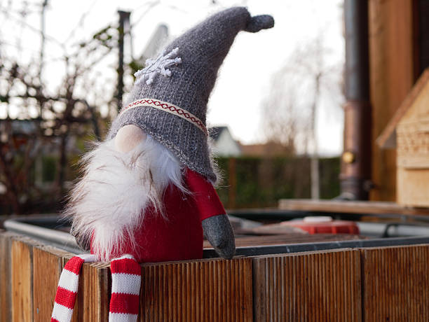 Handmade Christmas Gnome Doll with Long Decorative Legs Positioned on Wooden Structure in Outdoor Rustic Setting for Seasonal Holiday Imagery. High quality photo A festive Christmas gnome figurine with long striped legs sits on a wooden surface in a rustic outdoor environment. The gnome wears a gray hat with a snowflake and features fluffy white details. The cozy scene evokes holiday charm, winter decor, handmade crafts, and seasonal Christmas themes.