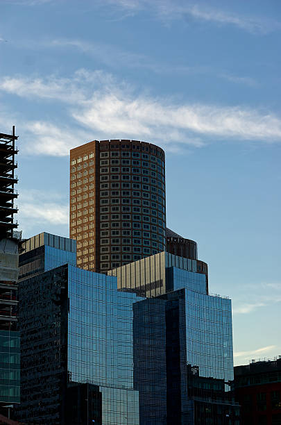 Downtown cityscape captures skyscrapers at dusk, including tall cylindrical building distinctive facade and large windows reflecting evening light. Scattered clouds highlight clear blue sky.Boston, MA