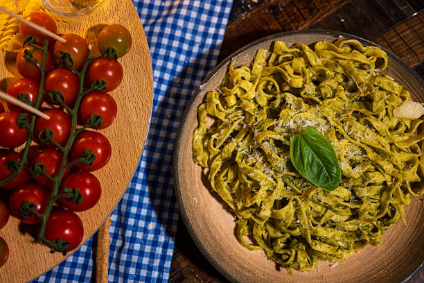 A top-view shot of a bowl of homemade pesto tagliatelle topped with grated parmesan and fresh basil. Bright cherry tomatoes on the vine sit beside the plate, creating a vibrant and rustic Italian dining atmosphere. Perfect for themes like Italian cuisine, fresh pasta, Mediterranean food, cooking, gourmet meals, and restaurant menus.