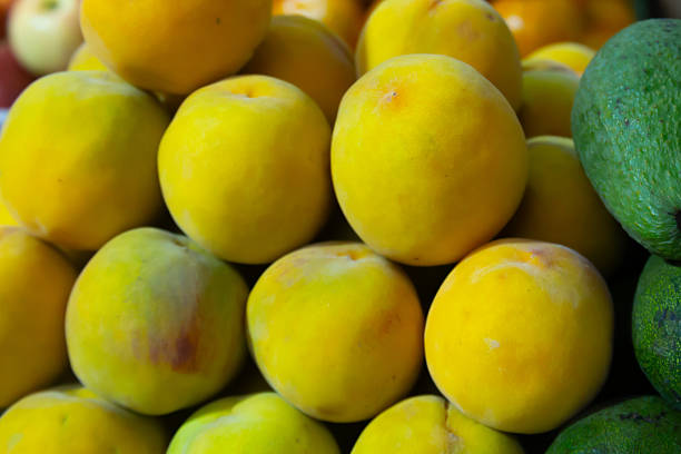 A pile of yellow peaches. The peaches are piled on top of each other. The pile is in front of a pile of green vegetables
