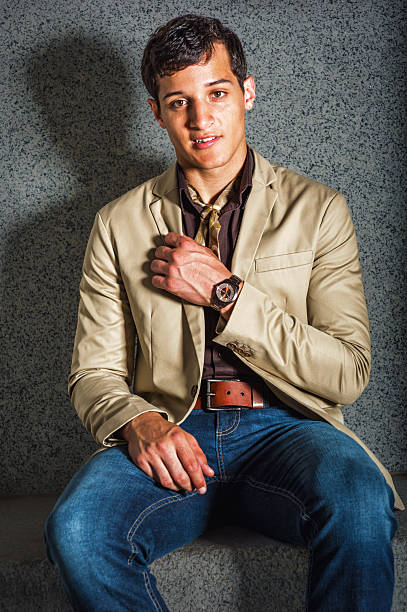 Stylish Young Man in Casual Attire. A young man sits confidently on a concrete bench. He wears a beige blazer, brown shirt, and blue jeans, accessorized with a watch.