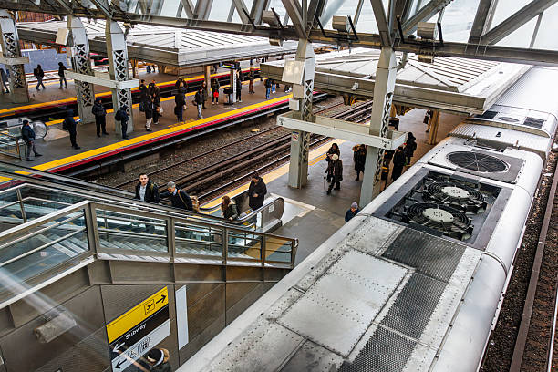 High angle view of a busy train station in Queens, New York City. Passengers wait on multiple platforms and use an escalator to move between levels. A commuter train is stopped at one of the tracks.