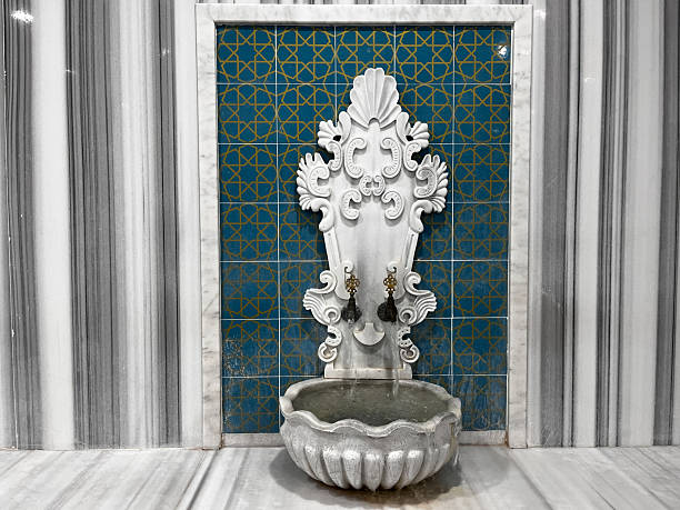 A white, carved stone fountain (kurna) with gold-leaf details, set against marble-patterned walls. Traditional Turkish bath architecture, tranquility, and history.