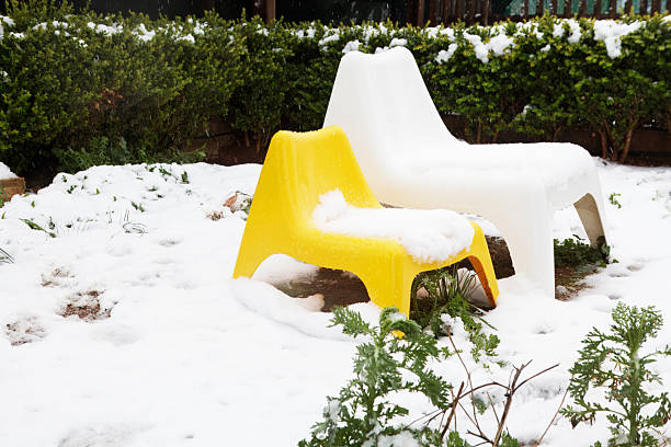 Two colorful plastic garden chairs, one yellow and one white, sit unused in a snowy backyard, partially covered in fresh snow with green bushes in the background. The playful summer furniture contrasted with the cold white snow creates a whimsical, nostalgic mood, perfect for concepts of changing seasons, unexpected weather, family life, holiday time, off-season tourism and outdoor living.
