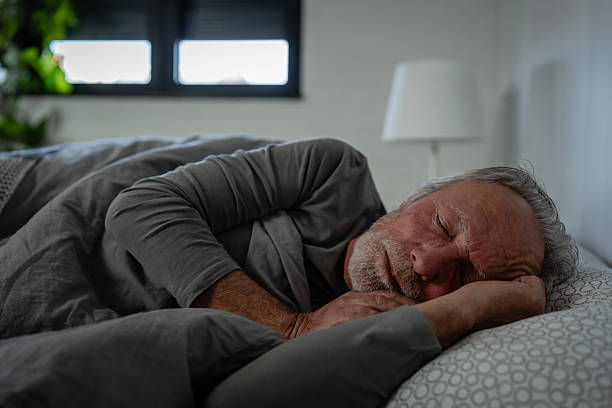 istock Elderly man sleeping peacefully in bed, resting on his side under a gray blanket in a softly lit bedroom 2250552749