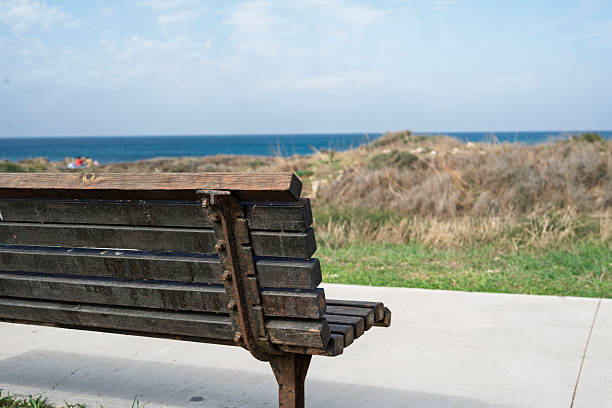 A rustic, weathered wooden bench sits near the shoreline, facing the open sea under a bright sky. Dry coastal vegetation and blue ocean water fill the background, creating a calm and contemplative seaside atmosphere perfect for themes of solitude and relaxation.