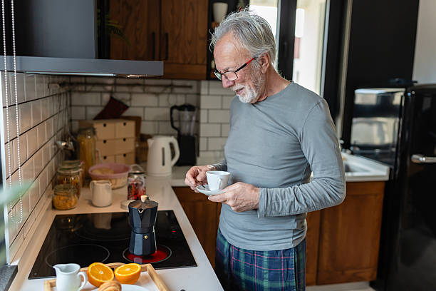 istock Smiling senior man preparing morning coffee in a cozy kitchen, holding a cup while waiting for a moka pot to brew beside fresh oranges 2250553932
