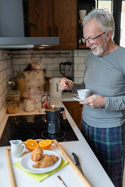 istock Smiling senior man preparing morning coffee in a cozy kitchen, holding a cup while waiting for a moka pot to brew beside fresh oranges 2250554073