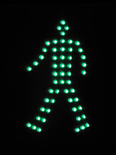 A green pedestrian traffic light signaling it is safe to walk. The light is mounted on a white pole with an urban background blurred out in soft focus.