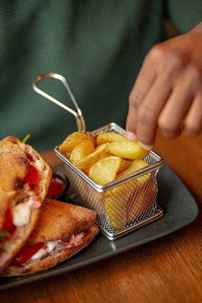 A neatly plated sandwich with fries sits on a wooden restaurant table, offering a casual lunch setting as a young Black man reaches for his fried potatoes.