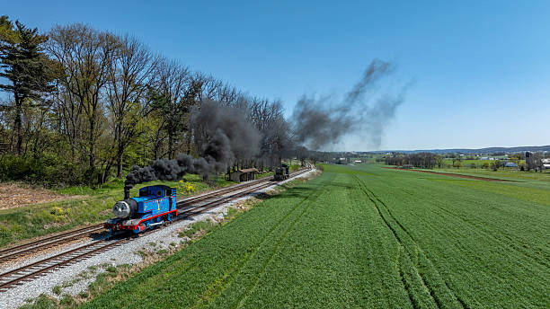 Ronks, Pennsylvania, April 17, 2025 - A blue steam train is traveling along a railway, emitting black smoke as it moves through lush green fields under a clear blue sky. Trees line the background, creating a serene scene.