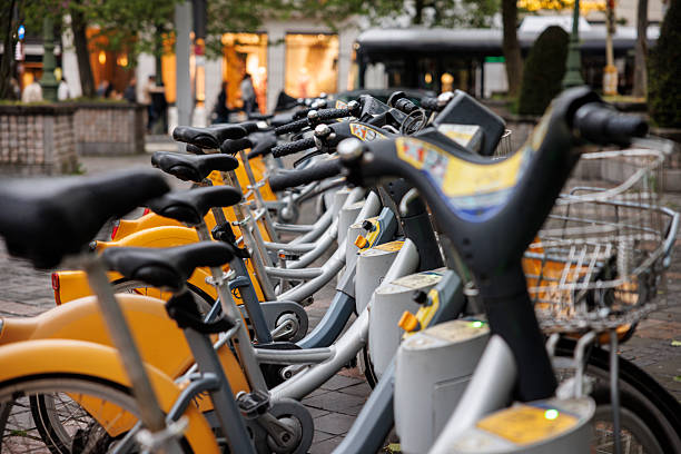 A row of classic, non-electric bicycles is parked at a public bike-sharing station in Brussels, Belgium. The yellow fenders and sturdy frames form a repeating visual rhythm, while soft evening light from nearby shop windows adds warmth to the scene. The bicycles are docked in place, ready for commuters and city travelers. Subtle reflections on the pavement and blurred movement in the background create a sense of urban atmosphere. This image captures sustainable mobility, everyday city life, and the practical convenience of bike-sharing systems widely used throughout European capitals.