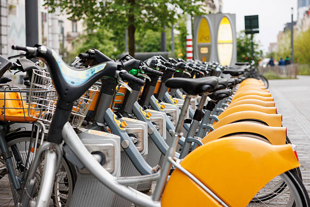 A long row of classic, non-electric bicycles is lined up at a public bike-sharing station in Brussels, Belgium. Their yellow fenders, metal baskets, and sturdy frames create a strong repeating pattern that reflects the structure and efficiency of the city's mobility system. Soft daylight illuminates the bicycles and the surrounding pavement, while trees and blurred urban elements in the background add depth and context. The image highlights sustainable transportation, urban commuting habits, and the role of bike-sharing programs in modern European cities.