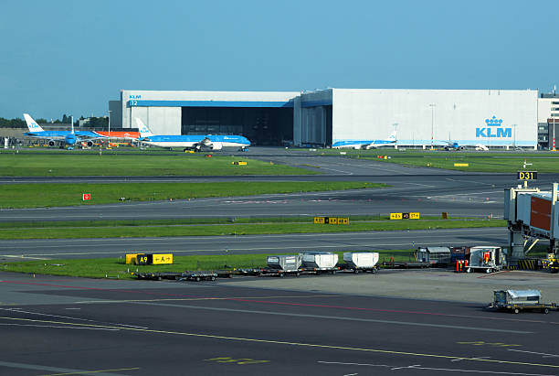 Haarlemmermeet, North Holland, Netherlands: exterior view of the KLM Engineering & Maintenance (E&M) division at Amsterdam Airport Schiphol (AMS), Netherlands. The image highlights the massive Hangar 12 facility, a central hub for airframe maintenance, repair, and overhaul (MRO) services. Multiple commercial aircraft are visible parked at the hangar bays undergoing technical engineering works, demonstrating the large-scale operational infrastructure and technical capacity of the airline's primary maintenance base.