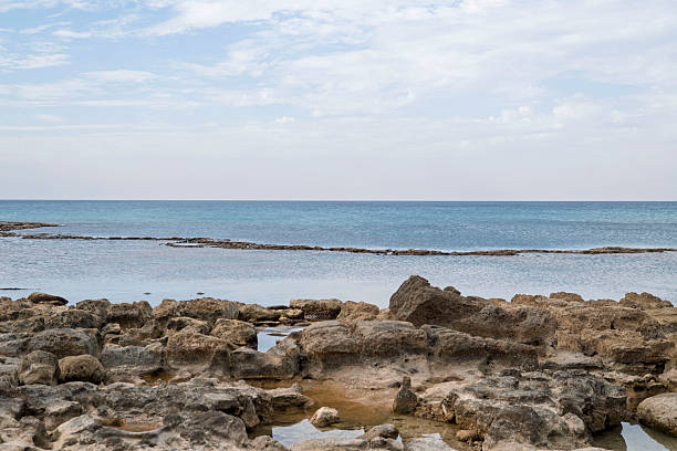 A wide coastal scene featuring jagged, exposed Kurkar (sandstone) reefs in the immediate foreground, leading to a long, low line of rock shelf that separates the shallow tide pool from the deep blue Mediterranean Sea horizon.