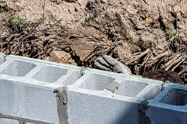 An anonymous worker's gloved hand and curly hair are just visible over the outside edge of a new home improvement in-law apartment addition construction site basement concrete block masonry wall with a straight, taut chalk line stretched above to ensure the wall is level. The cement is drying, but still quite wet as work continues.