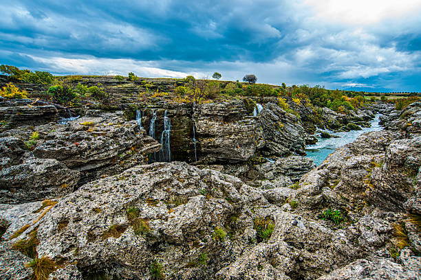 Thin waterfalls descend between layered limestone rocks into a narrow turquoise channel, surrounded by rugged stone, low shrubs, autumn vegetation, and a dramatic cloudy sky overhead above..