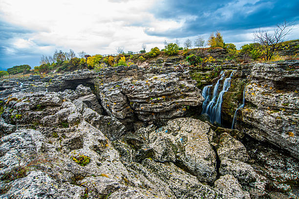 Thin waterfalls descend through deep gaps in layered limestone cliffs, surrounded by rugged rock blocks, sparse autumn shrubs, scattered trees on the ridge, and a bright, cloudy sky above.