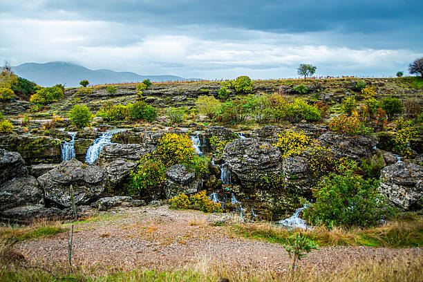 Multiple small waterfalls cascade over layered limestone terraces amid autumn shrubs and trees, with rocky ground in the foreground, green hills beyond, and heavy clouds hanging over distant mountains.