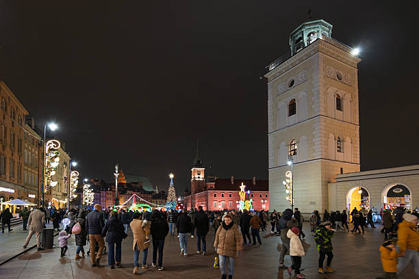 The historic Castle Square (Plac Zamkowy) in Warsaw, Poland, is packed with people at night. The Royal Castle clock tower overlooks a brightly lit Christmas tree and market area.