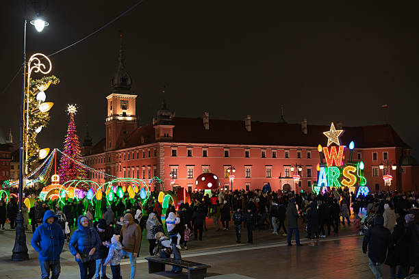 Wide view of a public square in Warsaw, Poland, featuring the Royal Castle and extensive Christmas light displays. A large crowd is gathered to enjoy the festive winter evening.