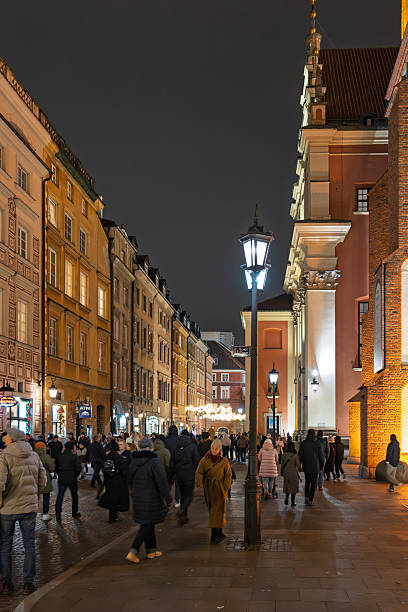 A busy, vertical view of a cobblestone street in Warsaw's Old Town at night, filled with crowds of pedestrians. The historic buildings are warmly illuminated by streetlights and shop windows.