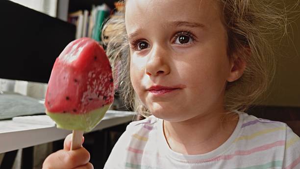 Cute happy little girl is eating ice cream in watermelon shape, while sitting carefree at home, illuminated by sunlight. Summer pleasures & happy childhood concept.