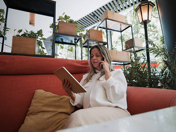 istock Businesswoman talking on smartphone while checking tablet on sofa in cafe 2250618362