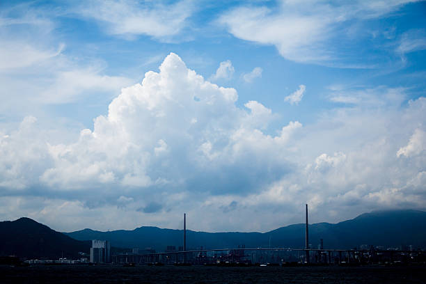 A scenic view of a suspension bridge with tall pylons, dramatic clouds, and a cityscape framed by mountains.
