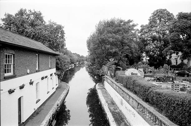 Grainy monochrome analog image capturing a serene canal reflecting trees and buildings, evoking a vintage vibe with its 35mm film aesthetic