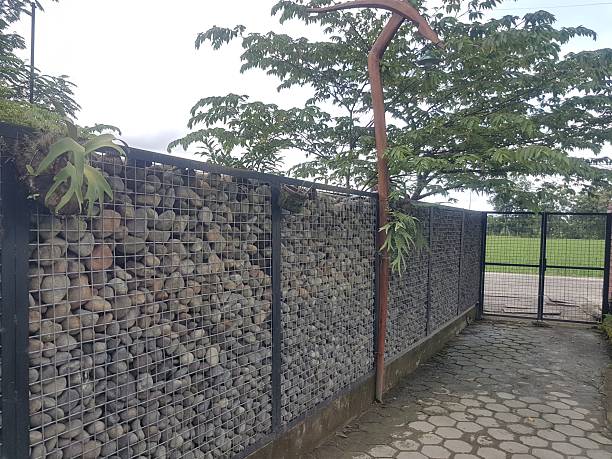 istock Gabion rock wall fence and metal gate next to a paved pathway. Unique landscaping design using stone filled wire mesh construction. Boundary and entrance to a green outdoor space. 2250638960