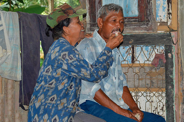 A heartwarming close-up of an elderly couple taking a break at their field hut, the man sits while his wife in a cap and jacket tenderly feeds him a piece of white snack or cake.