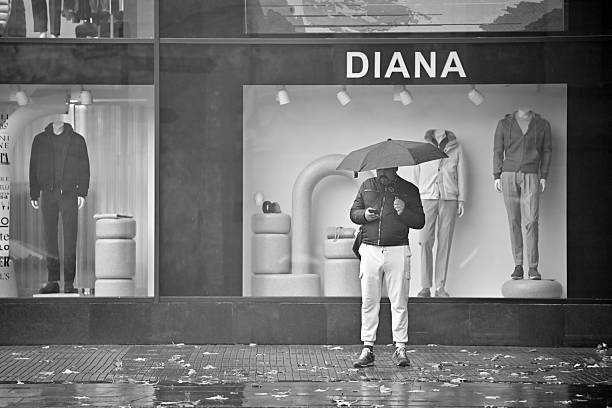 Yerevan Armenia 12.7.2025. A man is standing in the rain with an umbrella and smartphone, passing by a store window with a sign that says Diana