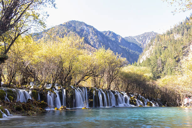 A turquoise mountain pool with cascading waterfalls, autumn trees and distant forested peaks creating serene atmosphere at Jiuzhaigou in Sichuan, China