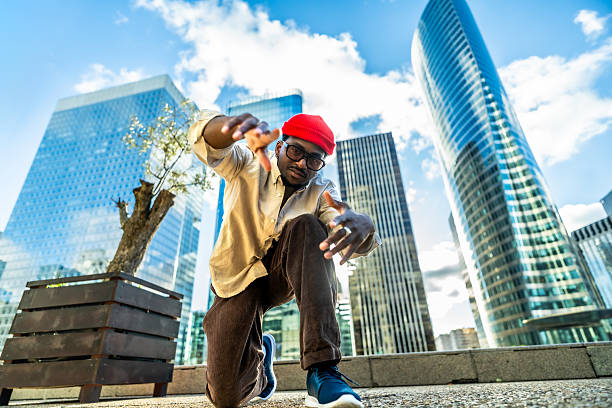 One young black man, wearing a red beanie, black glasses, and casual clothing, is squatting in an urban setting with modern skyscrapers reflecting blue sky, expressing confidence and street style