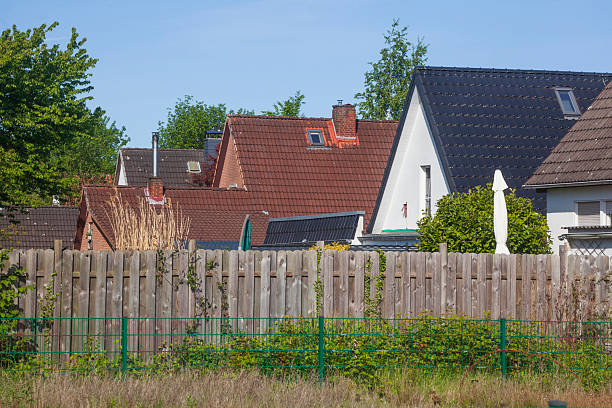 Amager, Copenhagen, Denmark - May 30th 2025:  A suburban house with solar panels on the roof, partially obscured by a neatly trimmed green hedge. The red brick house features a sloped roof and a prominent chimney. In the foreground, two mature street trees stand along the sidewalk, framing the residential setting. The image is taken on Kongelundsvej in Amager, a suburban area near central Copenhagen. The partly cloudy sky and soft daylight contribute to the calm and environmentally conscious atmosphere of this neighborhood.