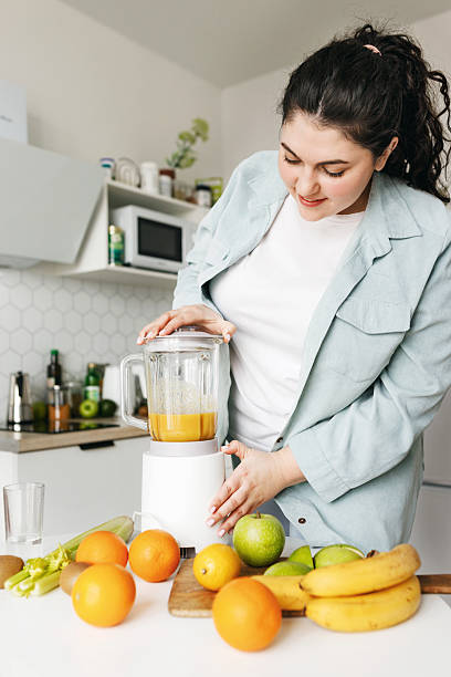 istock Healthy eating. Vertical image of plus size female in jacket at kitchen making smoothie from fruits using stationary blender for diet antioxidant breakfast before going to work in morning 2250669591