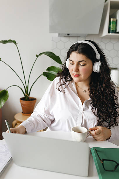 istock Vertical image of intelligent concentrated young Caucasian female using education platform studying online on laptop in headphones, making notes while listening to lecture or watching video tutorial 2250669698