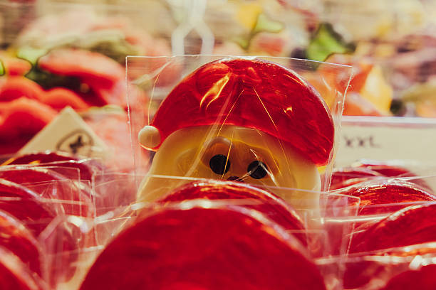 A close-up view of red and white lollipops shaped like Santa Claus, individually wrapped in clear plastic. The sweet treats are neatly arranged on a festive candy stand, surrounded by other colorful holiday confections. Warm lighting enhances the cheerful, seasonal mood of the image.
