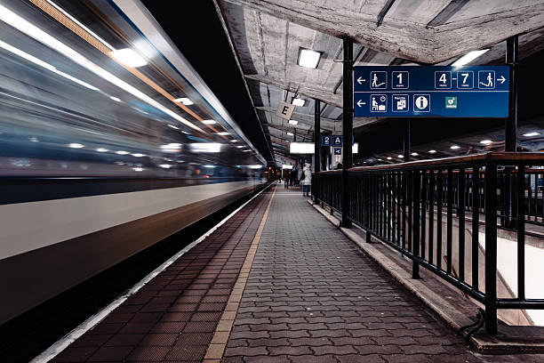 A fast-moving train blurs past a dimly lit platform at a concrete urban train station. Blue signage overhead directs passengers to various tracks and facilities, while a few bundled-up individuals wait in the cold. The long exposure emphasizes speed and the raw, utilitarian architecture of the location.