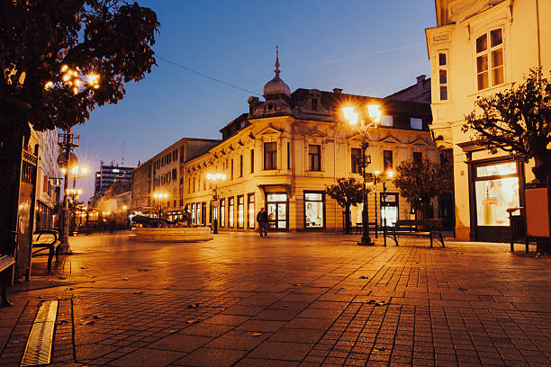 A quiet cobblestone pedestrian street glows under vintage-style street lamps at dusk, surrounded by historic European buildings and boutique shop windows. The deep blue sky contrasts beautifully with the warm, golden tones of the illuminated architecture, creating a peaceful urban ambiance.