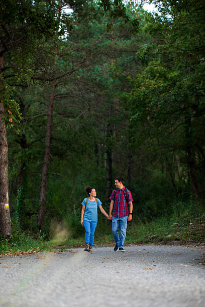 A smiling couple holds hands as they stroll along a gravel trail surrounded by tall trees and lush greenery.