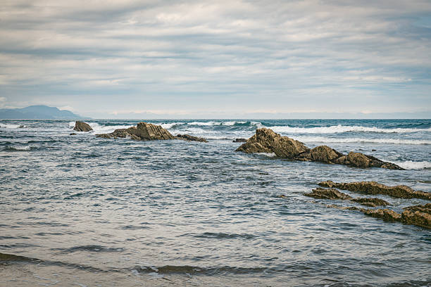 A tranquil coastal scene features a sandy beach, rugged rock formations, and a distant cliff. Gentle waves, zumaia beach spain,overcast skies, and calm mood evoke nature, exploration, and seaside serenity.