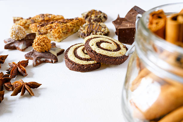Shallow-depth view of chocolate-vanilla spiral cookies surrounded by star anise, chocolate chunks, and the blurred foreground of a cinnamon-filled glass jar. The clean white background adds a modern feel while the spices create warmth and a homemade baking mood. Copy space available in the upper and side areas.