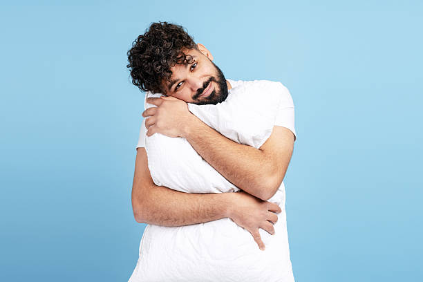 Young adult man enjoying hugging a soft pillow, relaxing and smiling on blue background