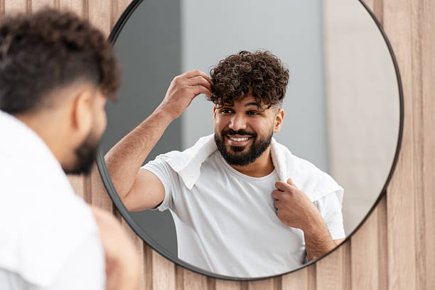Young adult man smiling, touching curly hair, looking at mirror in bathroom
