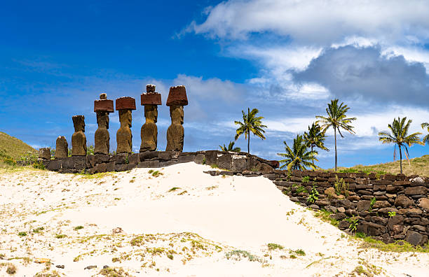 Moai statues with red pukao topknots stand on Ahu Nau Nau at Anakena Beach, Easter Island. White coral sand and palm trees are in the foreground