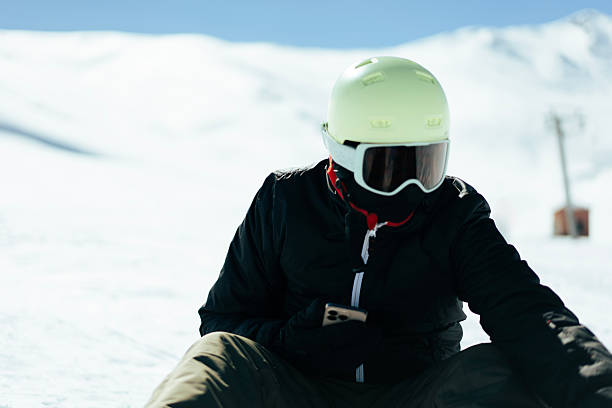 Snowboarder wearing helmet and goggles sitting on snowy mountain slope checking smartphone